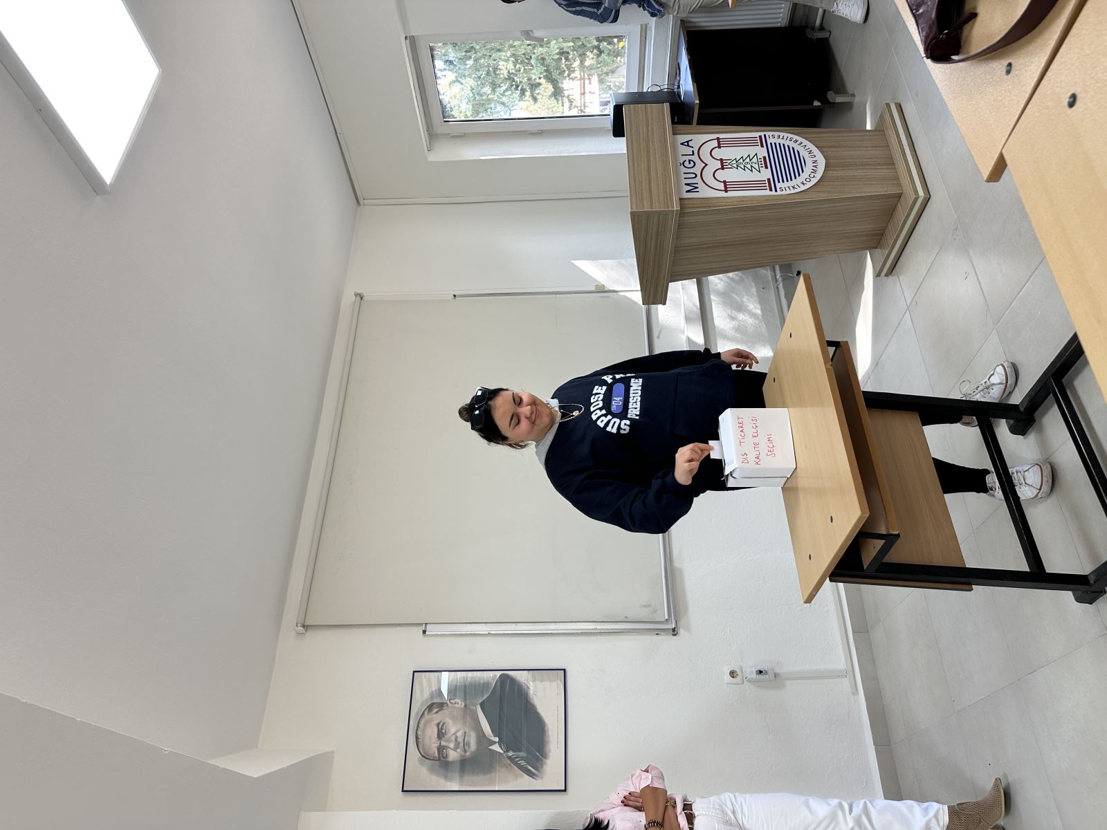 In the same classroom, a young woman is standing in front of a small desk, appearing to drop a piece of paper into a box labeled “Foreign Trade Quality Ambassador Election.” She is wearing a navy-blue sweatshirt and white sneakers, with her hair tied in a bun. On the right side, there is a wooden lectern with the Muğla Sıtkı Koçman University logo. On the left, a person wearing white pants and a pink shirt is standing with their arms crossed. The wall behind them features a portrait of Atatürk